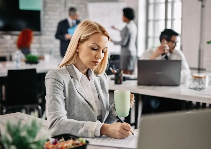 Professional woman writing notes at desk with coffee in open-plan office Business directory